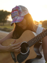 Person wearing Born In Earth Pink Trucker Hat playing an acoustic guitar with a sunset background