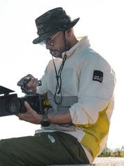 Person holding a camera, wearing Camp Khaki Safari Hat and a white jacket with a yellow patch.