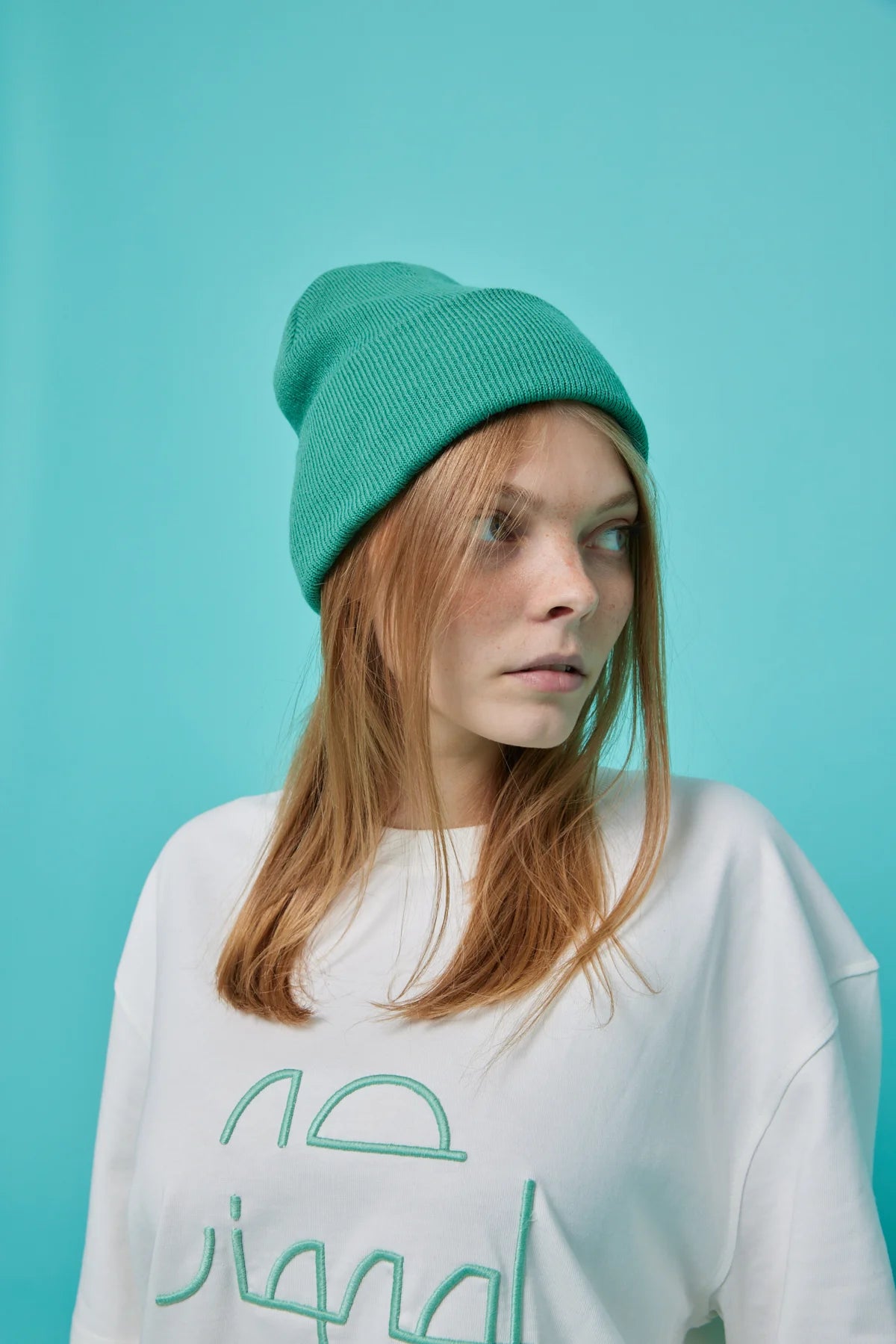 Person wearing a white sweatshirt with green text and a green beanie against a teal background