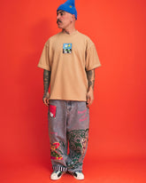 Person wearing a beige t-shirt with a graphic design and gray pants with colorful patches on an orange background