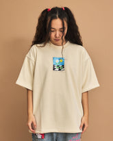 Person wearing a beige oversized t-shirt with a graphic design on a brown background