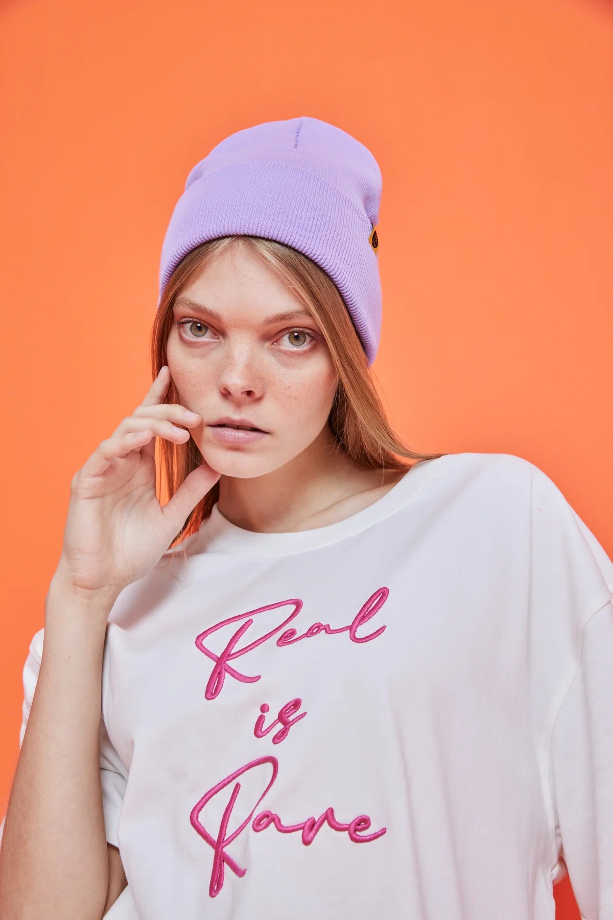 Woman wearing a purple beanie and white t-shirt with 'Real is Rare' text on an orange background