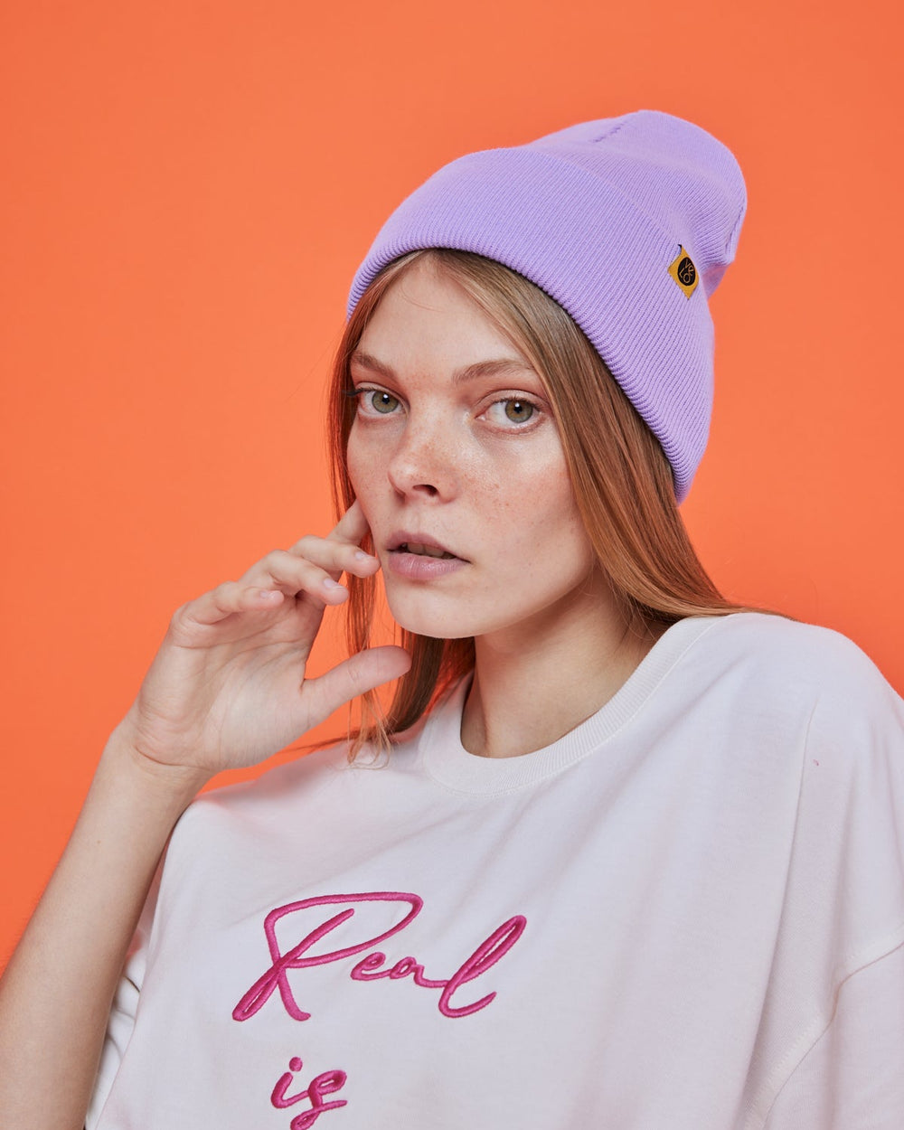 Person wearing a lilac purple beanie and white shirt with pink text against an orange background
