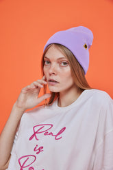 Person wearing a lilac purple beanie and white shirt with pink text against an orange background
