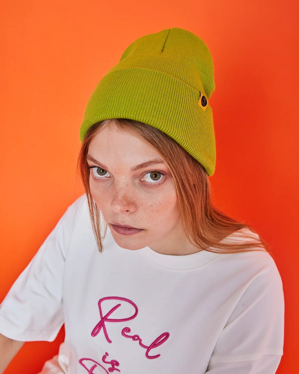 Person wearing a green beanie and white t-shirt with pink text on an orange background