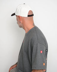 Man wearing a gray t-shirt with visible brand logos on a white background and showing brown flip tag of the cap