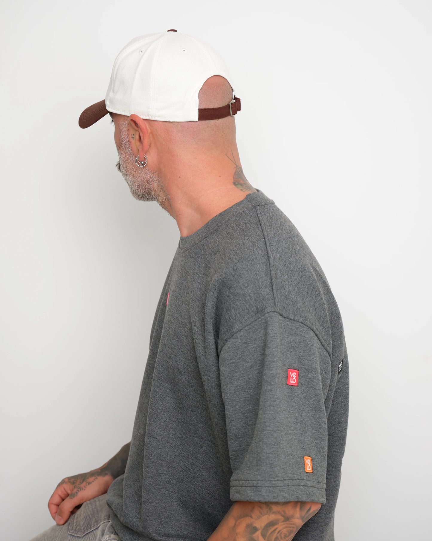 side look of man wearing a gray t-shirt with a small velo logos stitched and a white baseball cap with brown brim showing brown flip tag.