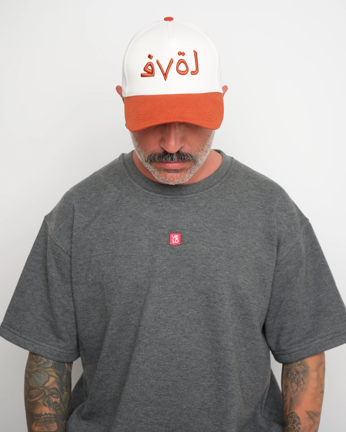 Man wearing a gray t-shirt with a small red logo and a white baseball cap with an orange brim and orange Arabic text on a plain background.