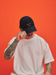 Person wearing a navy blue corduroy cap with text and a white t-shirt against an orange background