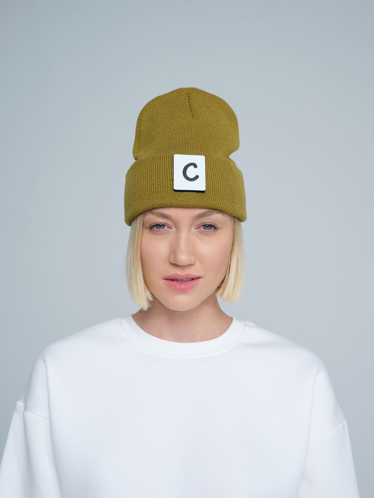 A person wearing olive beanie with 'C' letter patch attached on front on a light gray background