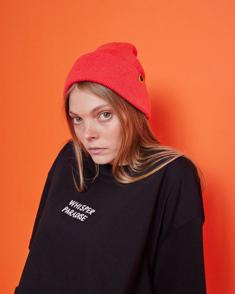 Person wearing a black oversized t-shirt with text and a red beanie against an orange background