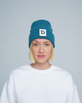 A person wearing teal beanie with 'D' letter patch attached on the front on a light gray background