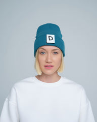 A person wearing teal beanie with 'D' letter patch attached on the front on a light gray background