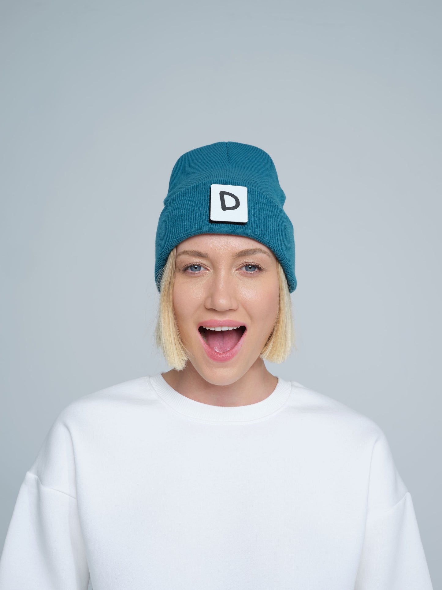 A person wearing teal beanie with 'D' letter patch attached on the front on a light gray background