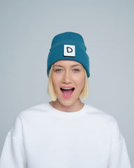 A person wearing teal beanie with 'D' letter patch attached on the front on a light gray background