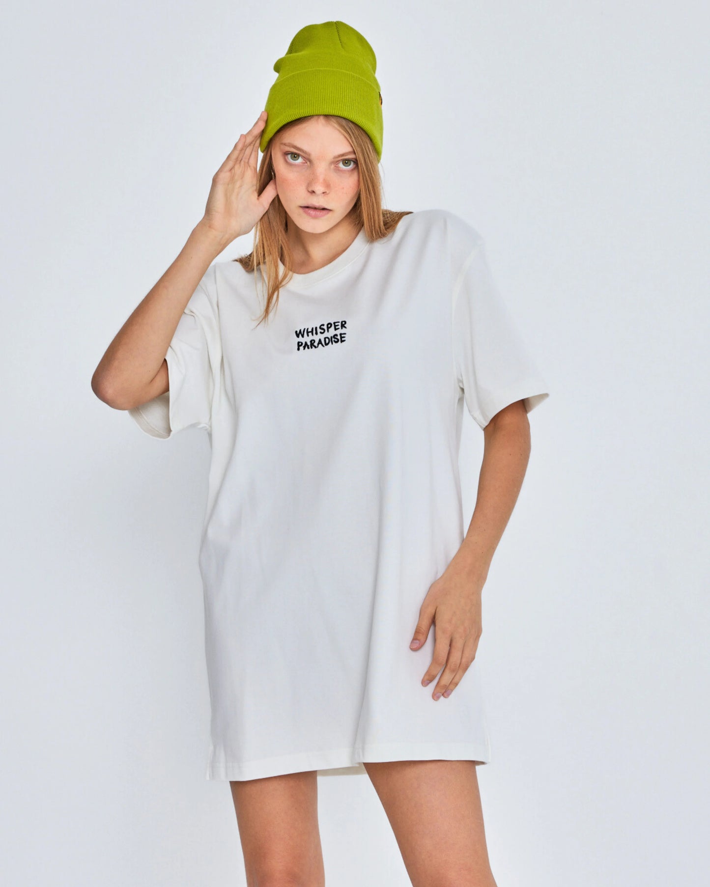 Person wearing a white oversized t-shirt with text and a green beanie on a plain background