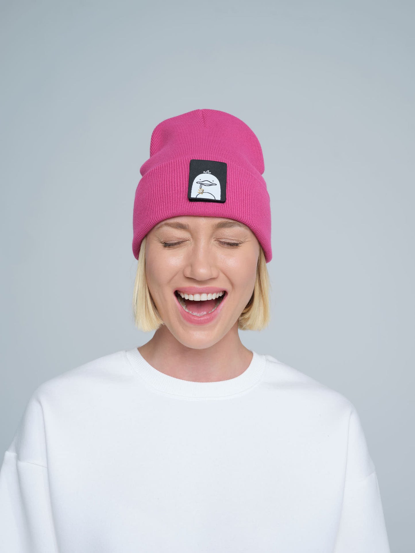 A person wearing a pink beanie with a 'duck' animal design on the fold, part of a white crew neck sweater.