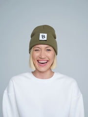 A person wearing an khaki beanie hat with a 'B' design patch on it, smiling against a white background.
