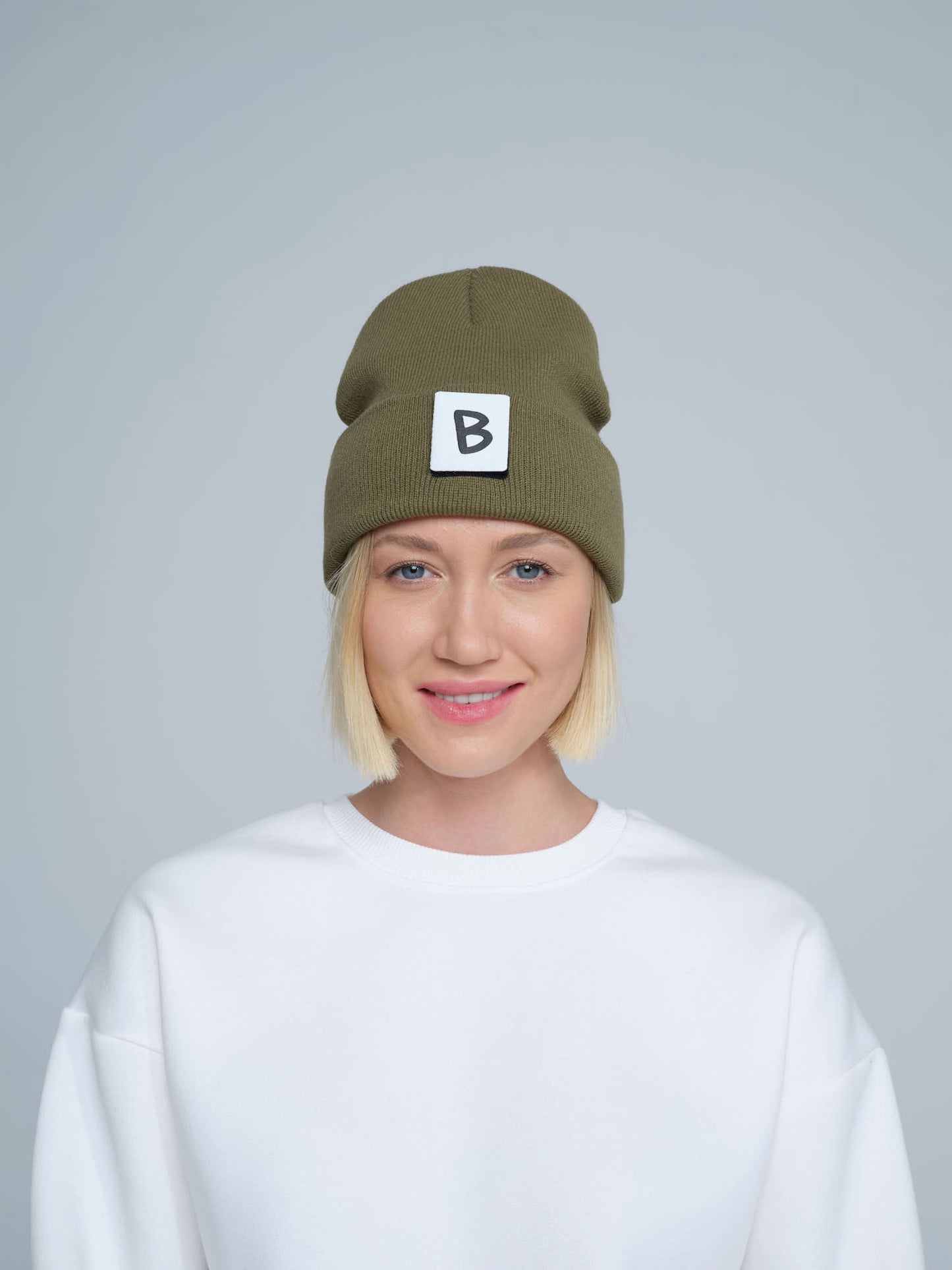 A person wearing an olive green beanie hat with a 'B' design patch on it, smiling against a white background.