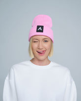 A person wearing a pink beanie with a design on top and the letter 'A' on it, paired with a white crew neck sweater.