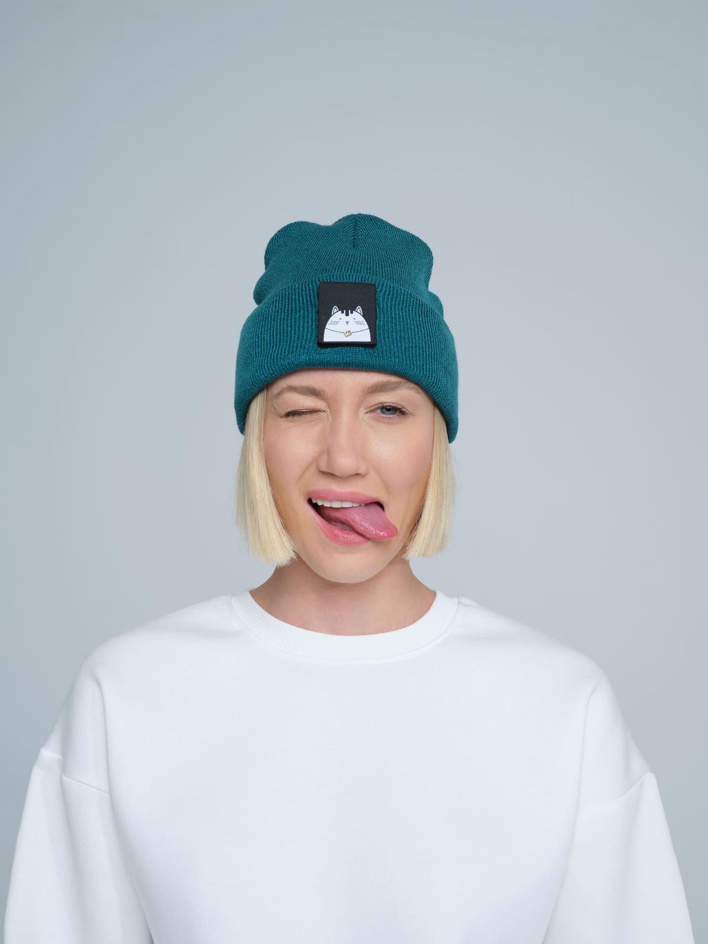 A female wearing a teal green beanie hat with a cat design on the cuff, making a funny face.