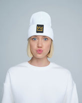 A person wearing a white beanie with a design and a crown symbol on it, standing in a casual pose.