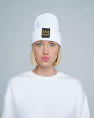 A person wearing a white beanie with a design and a crown symbol on it, standing in a casual pose.