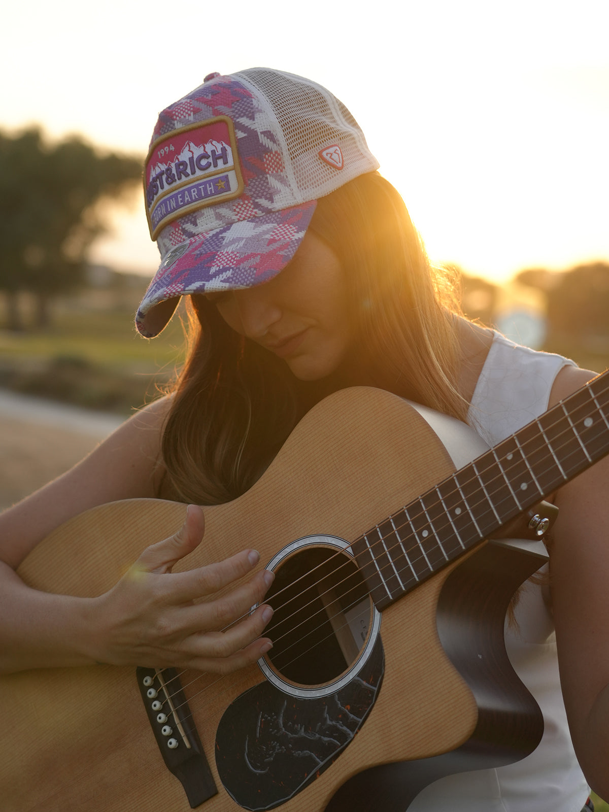 Person wearing Born In Earth Pink Trucker Hat playing an acoustic guitar with a sunset background