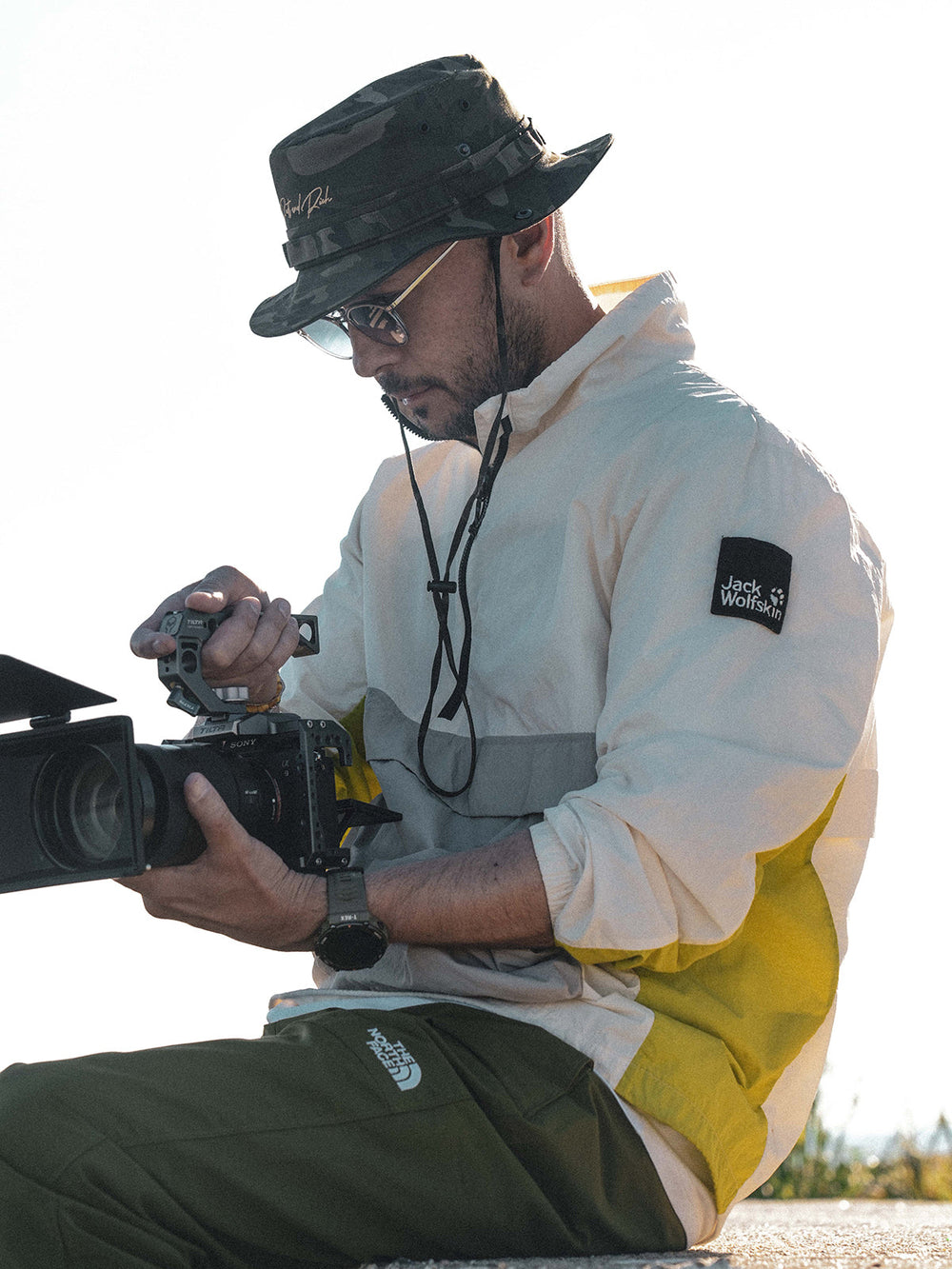 Person holding a camera, wearing Camp Khaki Safari Hat and a white jacket with a yellow patch.