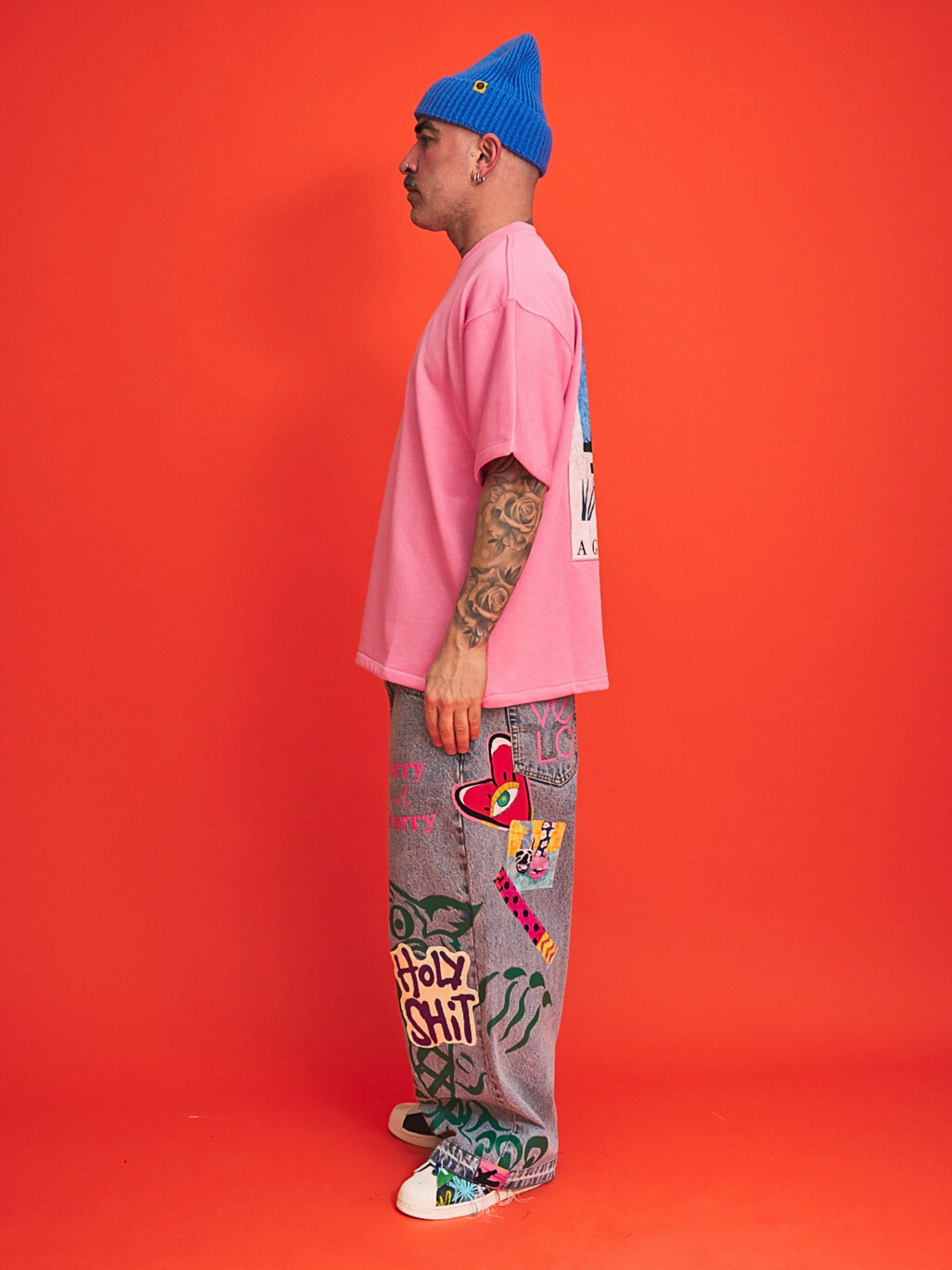 Person wearing a pink shirt, gray pants with graffiti, and blue beanie on an orange background
