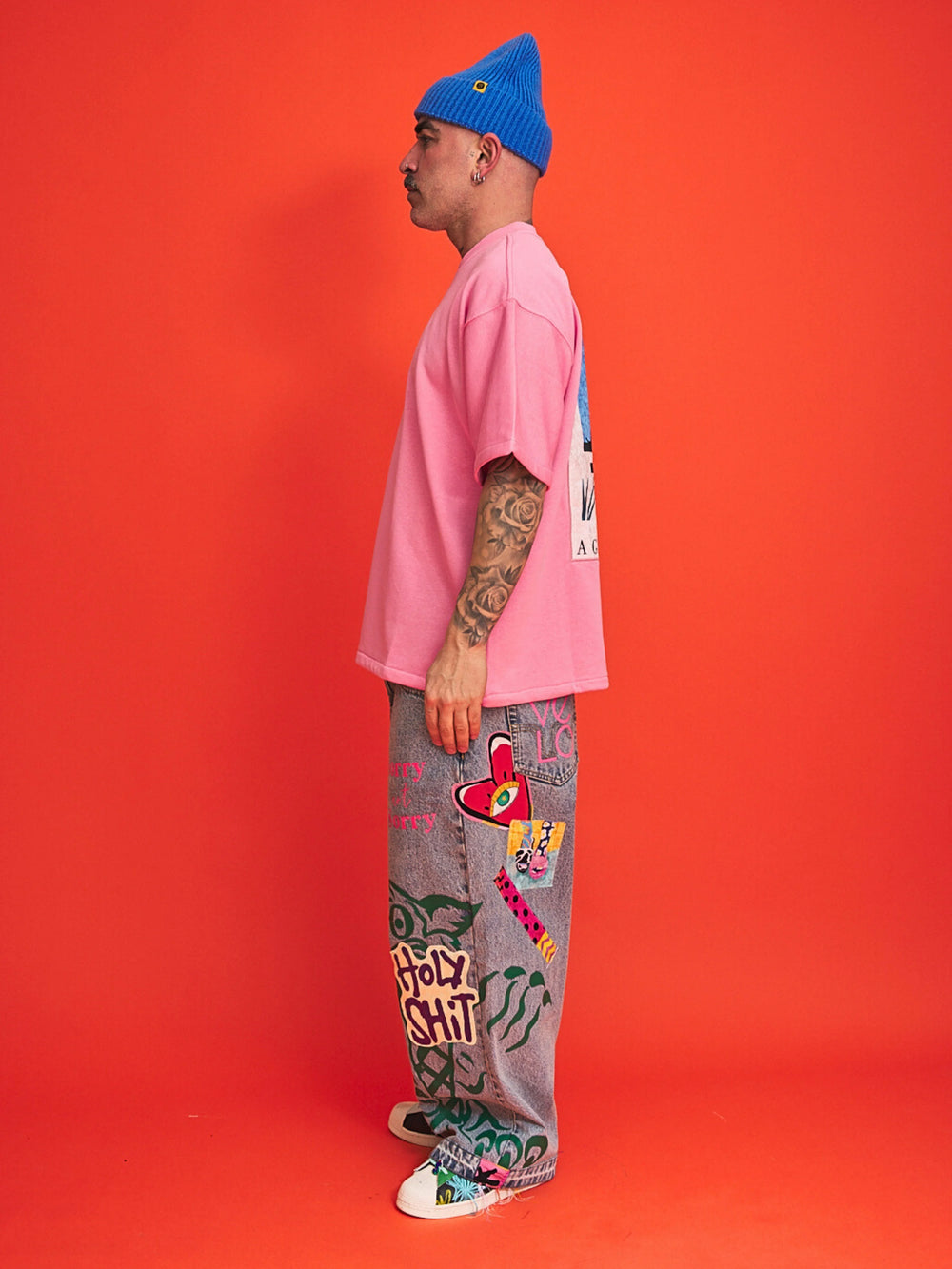 Person wearing a pink shirt, gray pants with graffiti, and blue beanie on an orange background