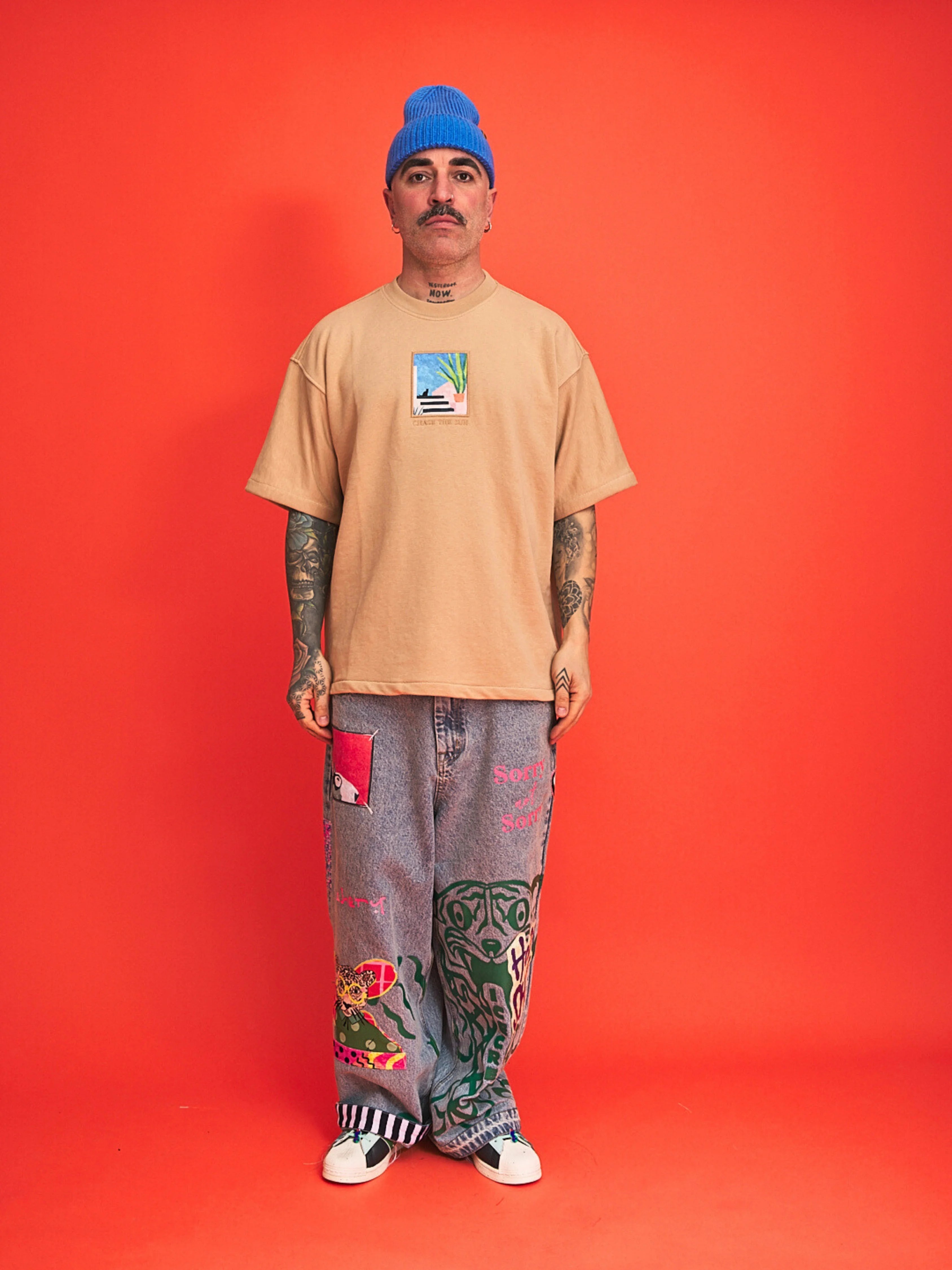 Man wearing a beige t-shirt, blue beanie, and patterned jeans on an orange background