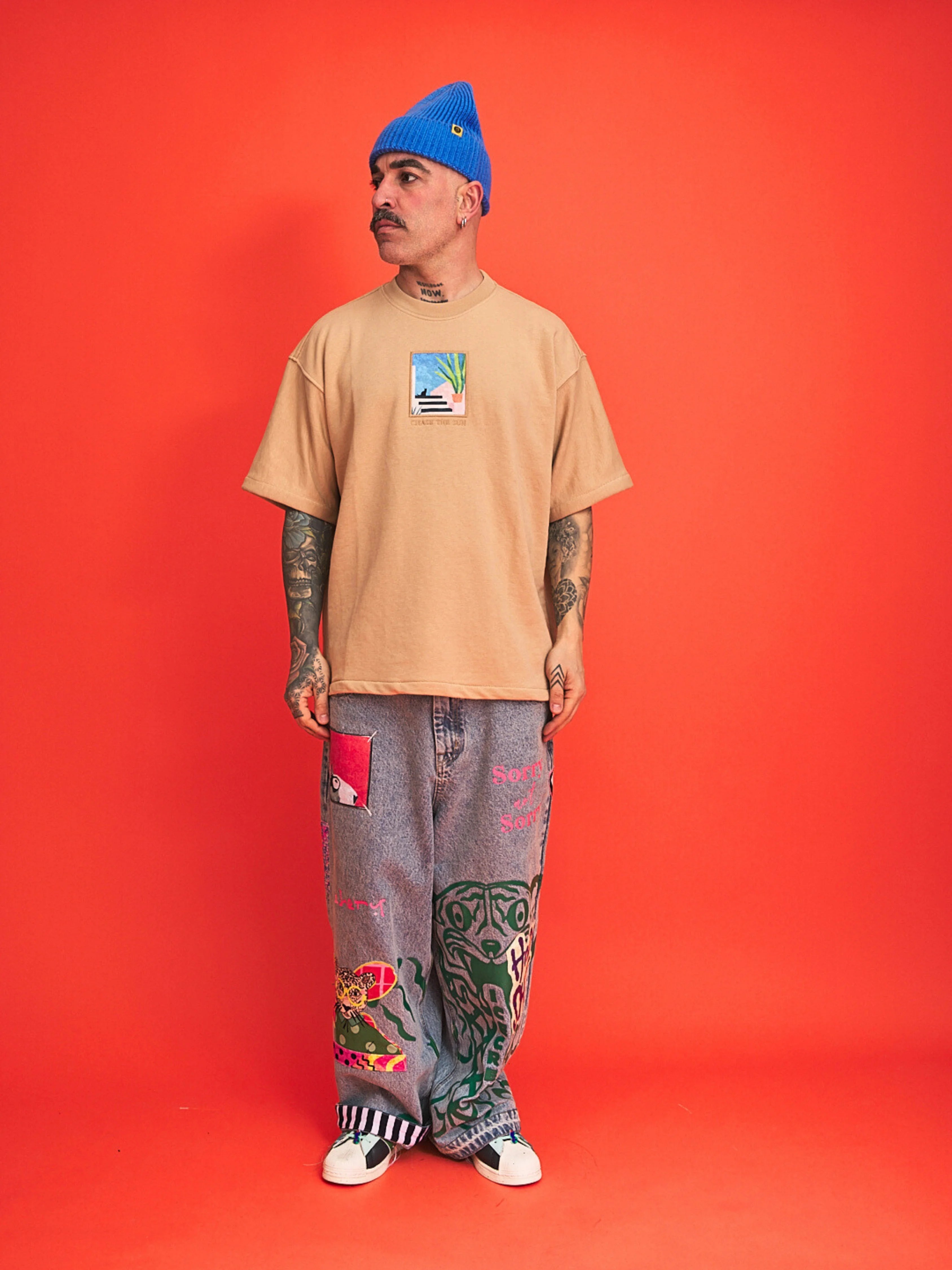 Man wearing a beige t-shirt, blue beanie, and patterned pants on an orange background