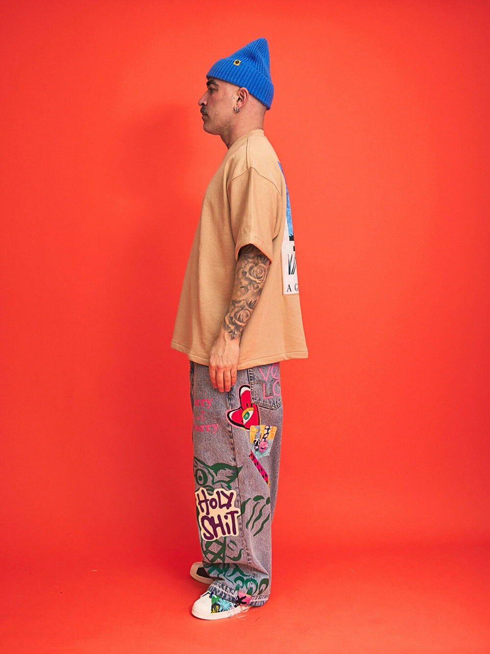 Person wearing a beige shirt, blue beanie, and colorful pants on an orange background
