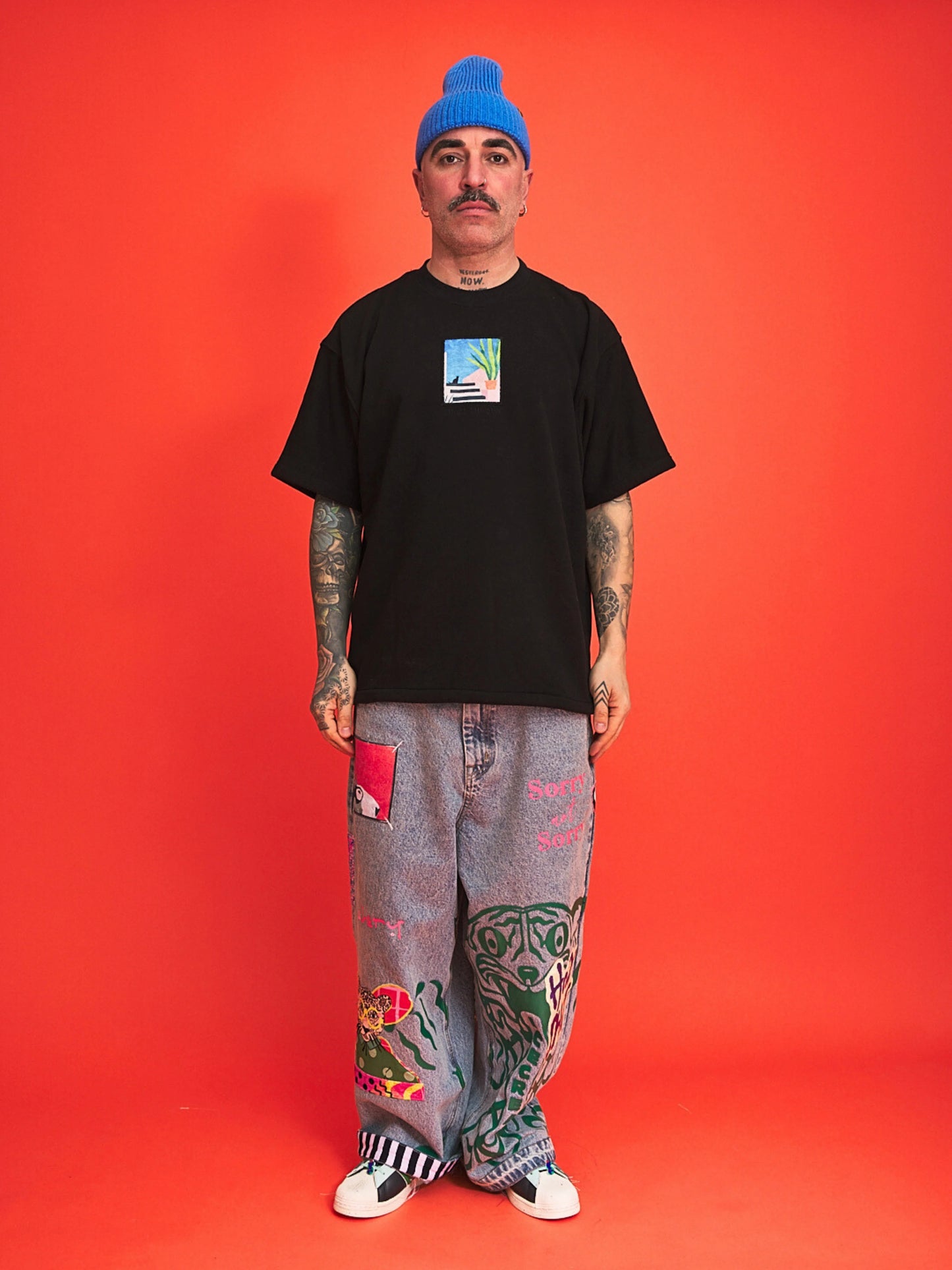 Person wearing a black t-shirt, blue beanie, and colorful pants on an orange background