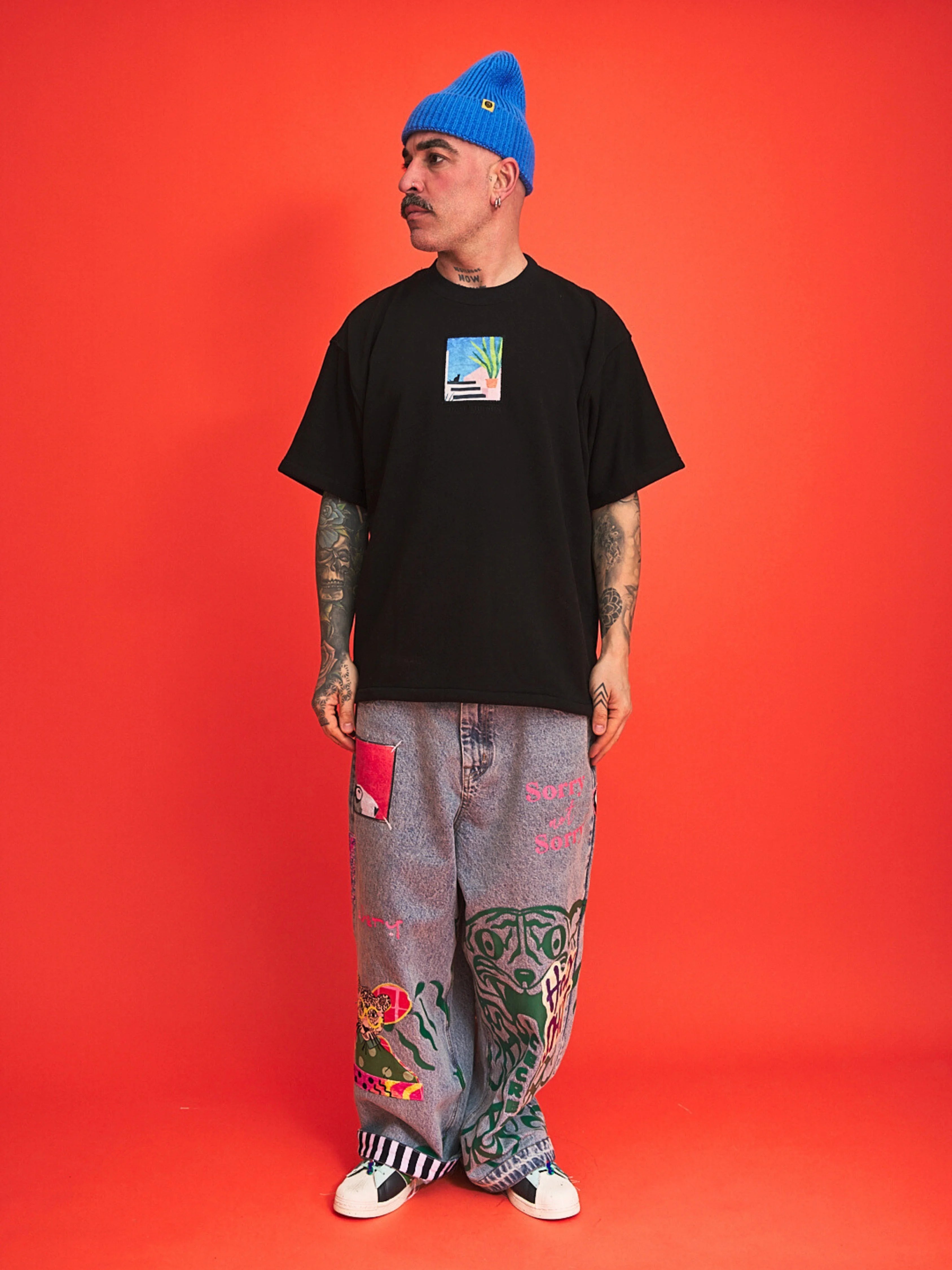 Person wearing a black t-shirt, blue beanie, and colorful pants on an orange background