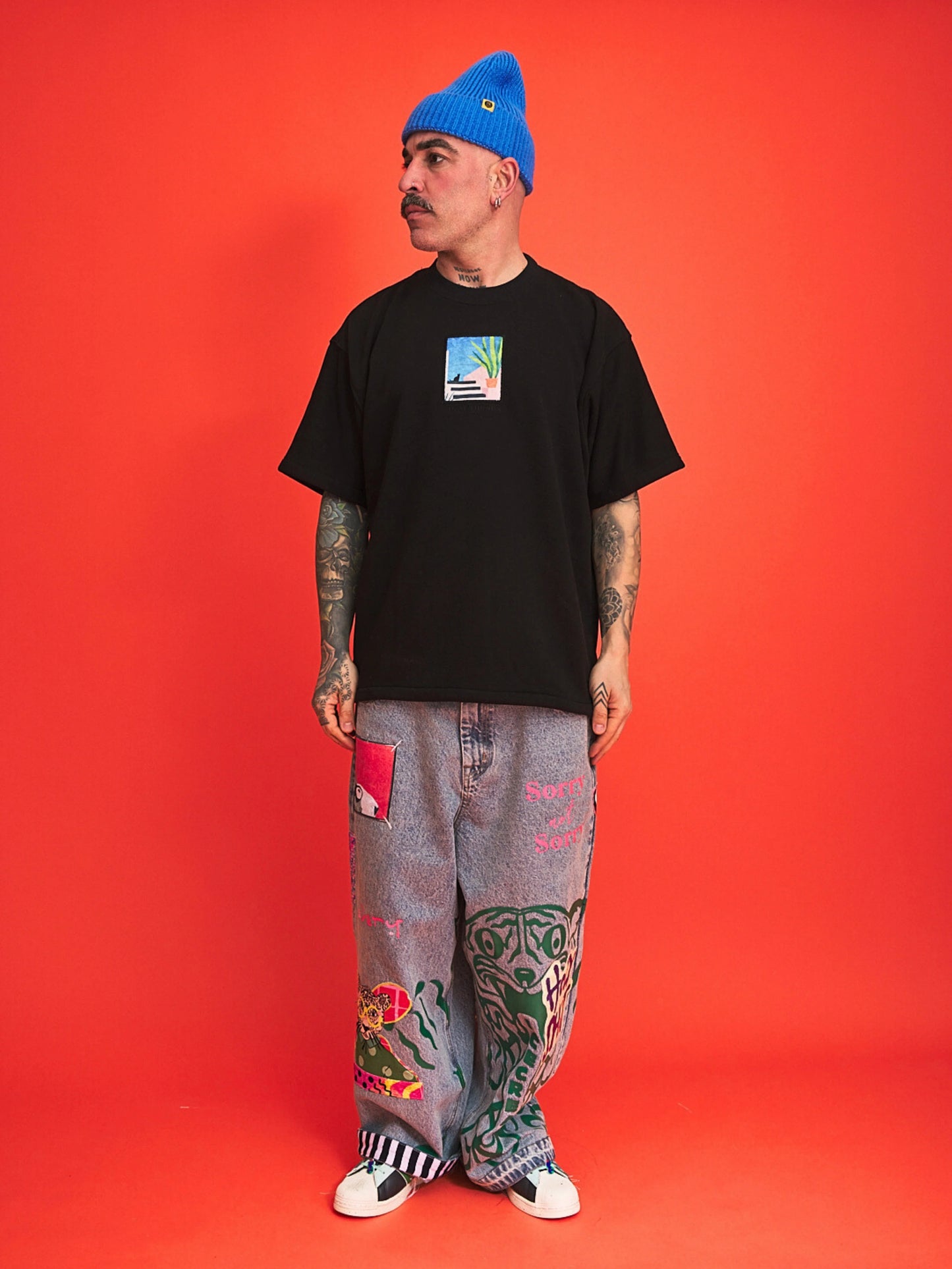 Person wearing a black t-shirt, blue beanie, and colorful pants on an orange background