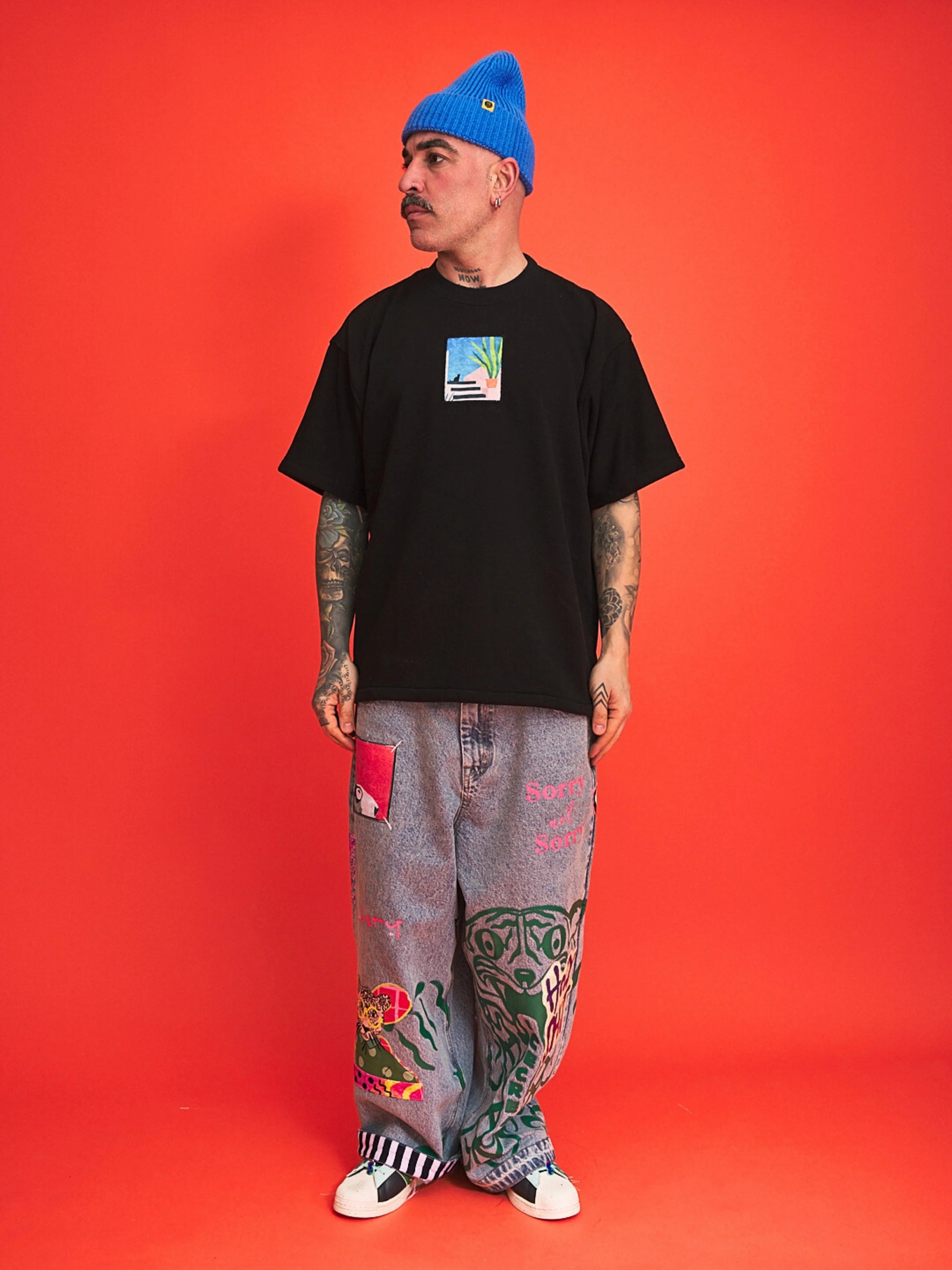 Person wearing a black t-shirt, blue beanie, and colorful pants on an orange background