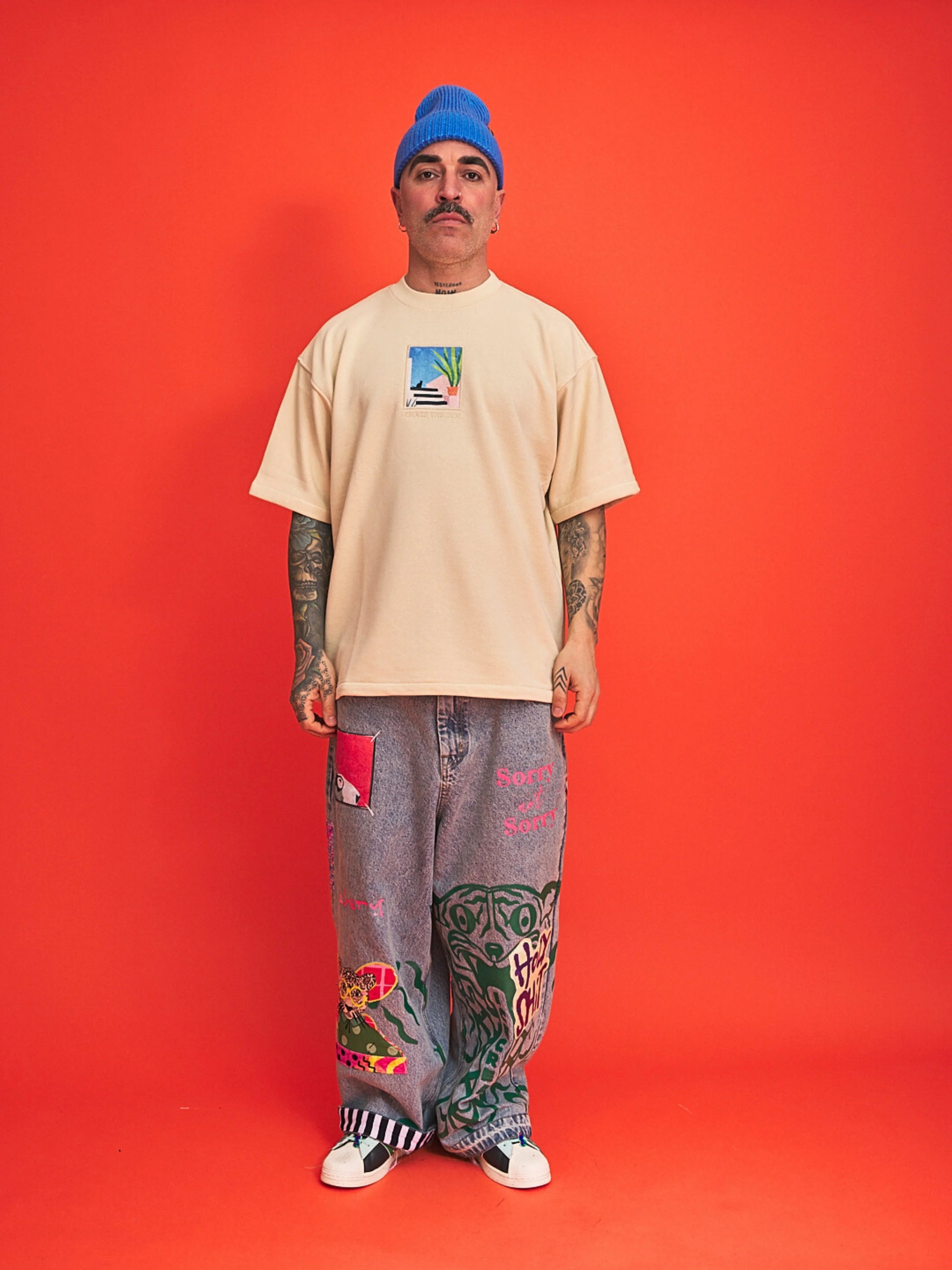 Person wearing a beige t-shirt, gray pants with designs, and a blue beanie on an orange background