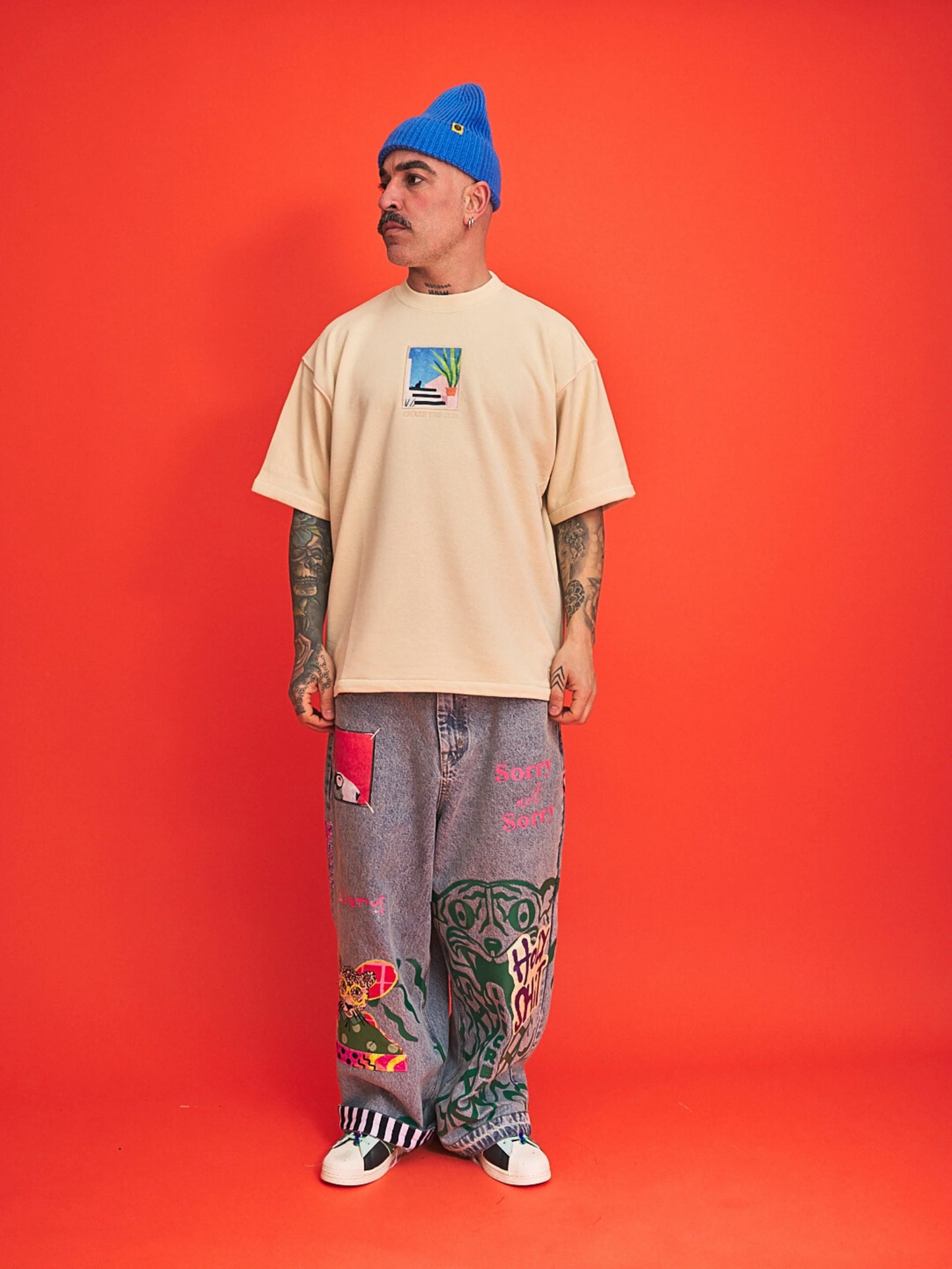 Man wearing a beige t-shirt, blue beanie, and patterned pants on an orange background