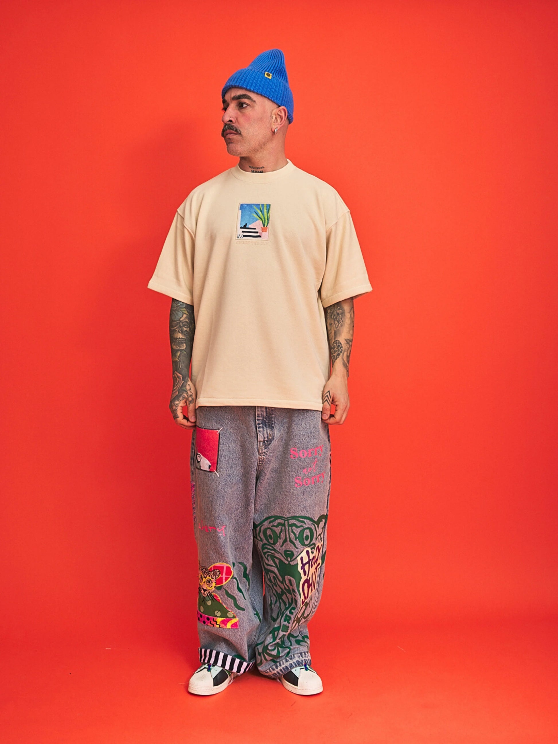 Man wearing a beige t-shirt, blue beanie, and patterned pants on an orange background