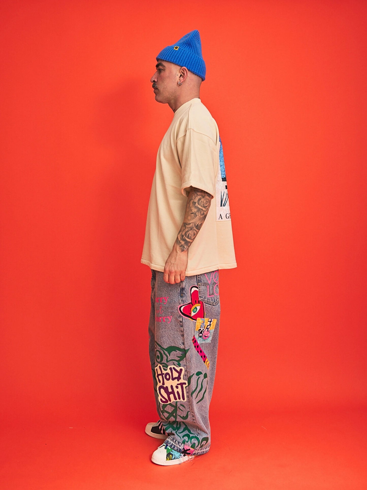 Person wearing a beige t-shirt, blue beanie, and colorful pants with text and graphics on an orange background