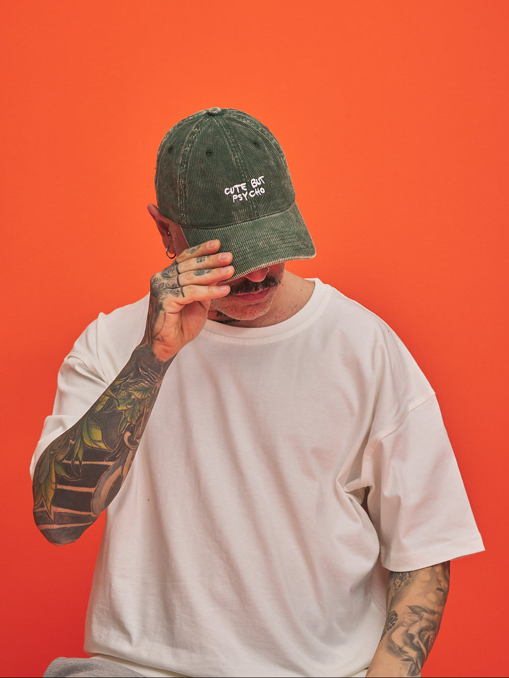 Person wearing a green corduroy cap with  'Cute But Psycho' text, covering their face against an orange background