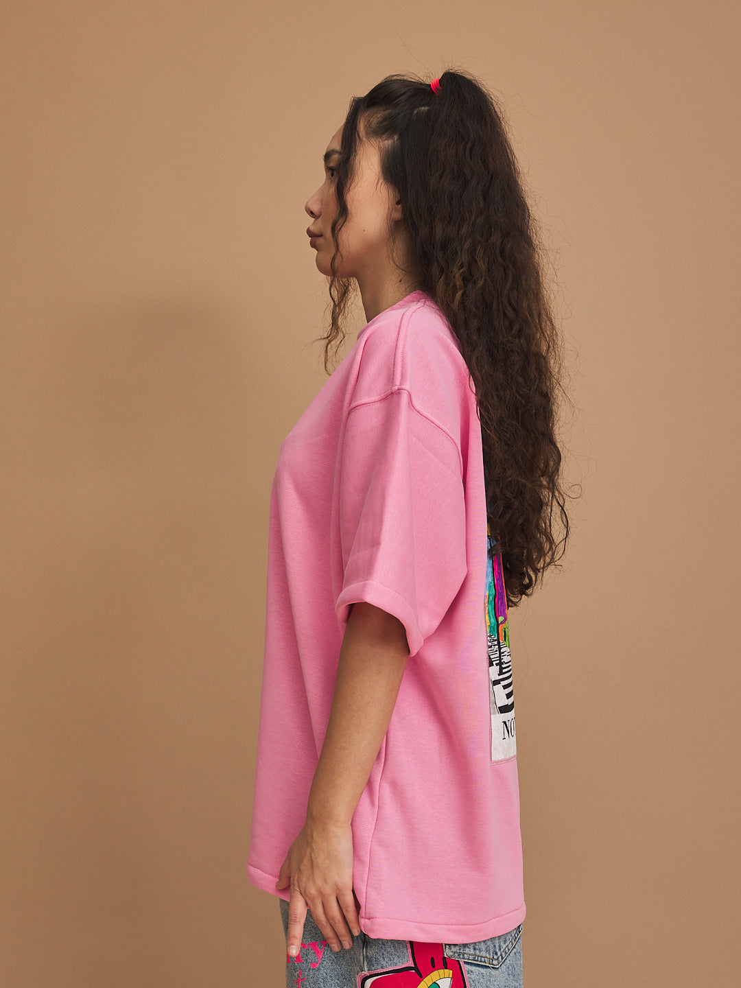 Person wearing a pink oversized t-shirt with colorful patches on a brown background