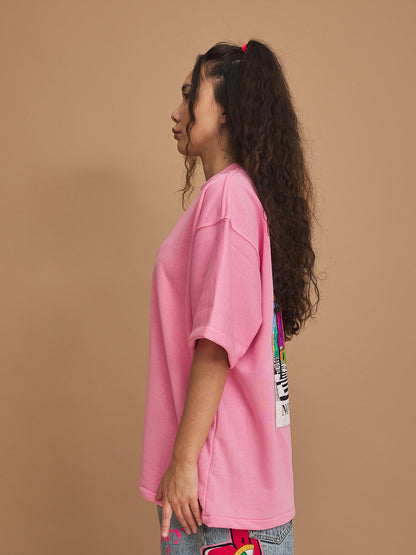 Person wearing a pink oversized t-shirt with colorful patches on a brown background