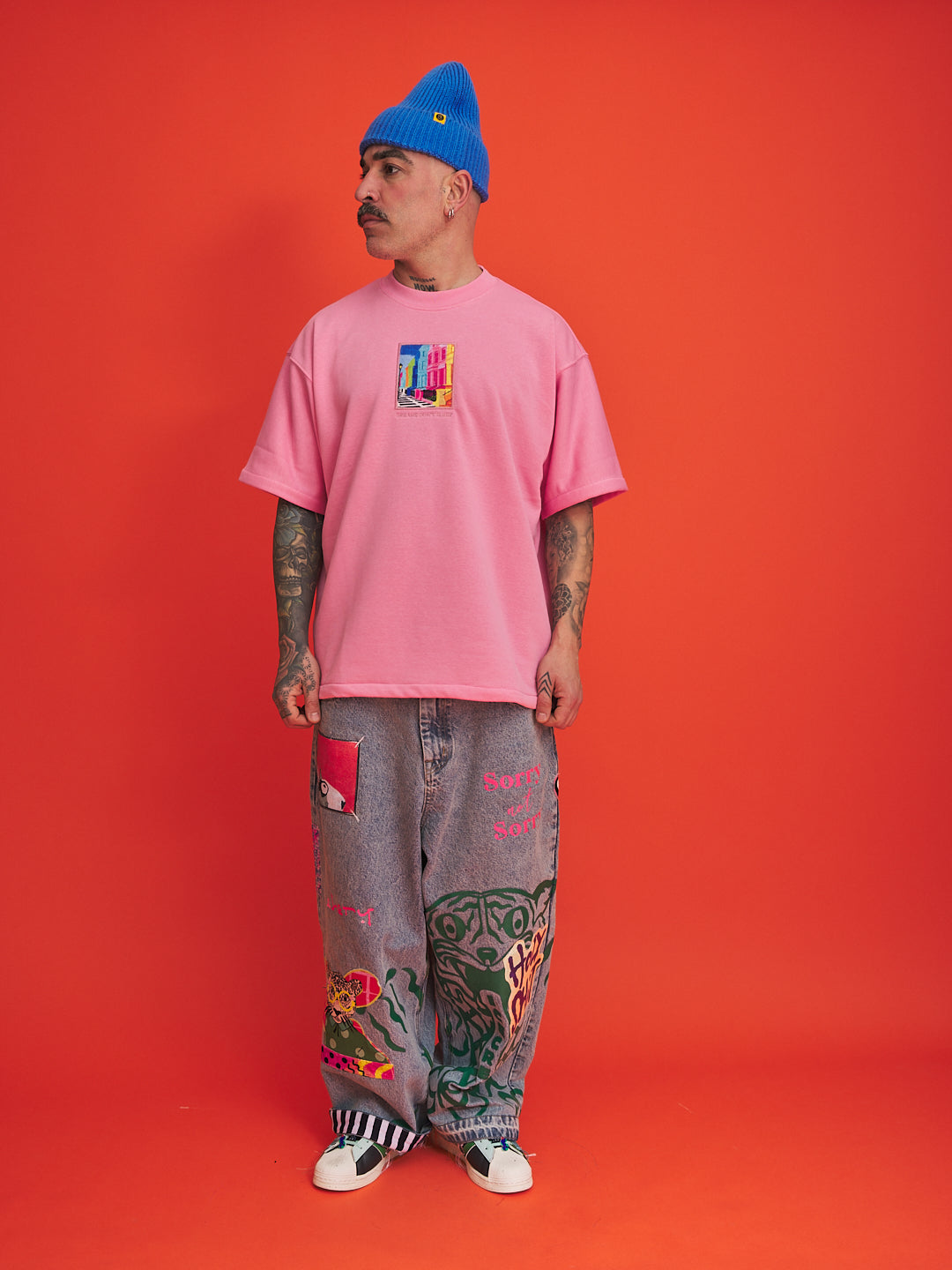 Person wearing a pink t-shirt and gray pants with tattoos, standing against an orange background