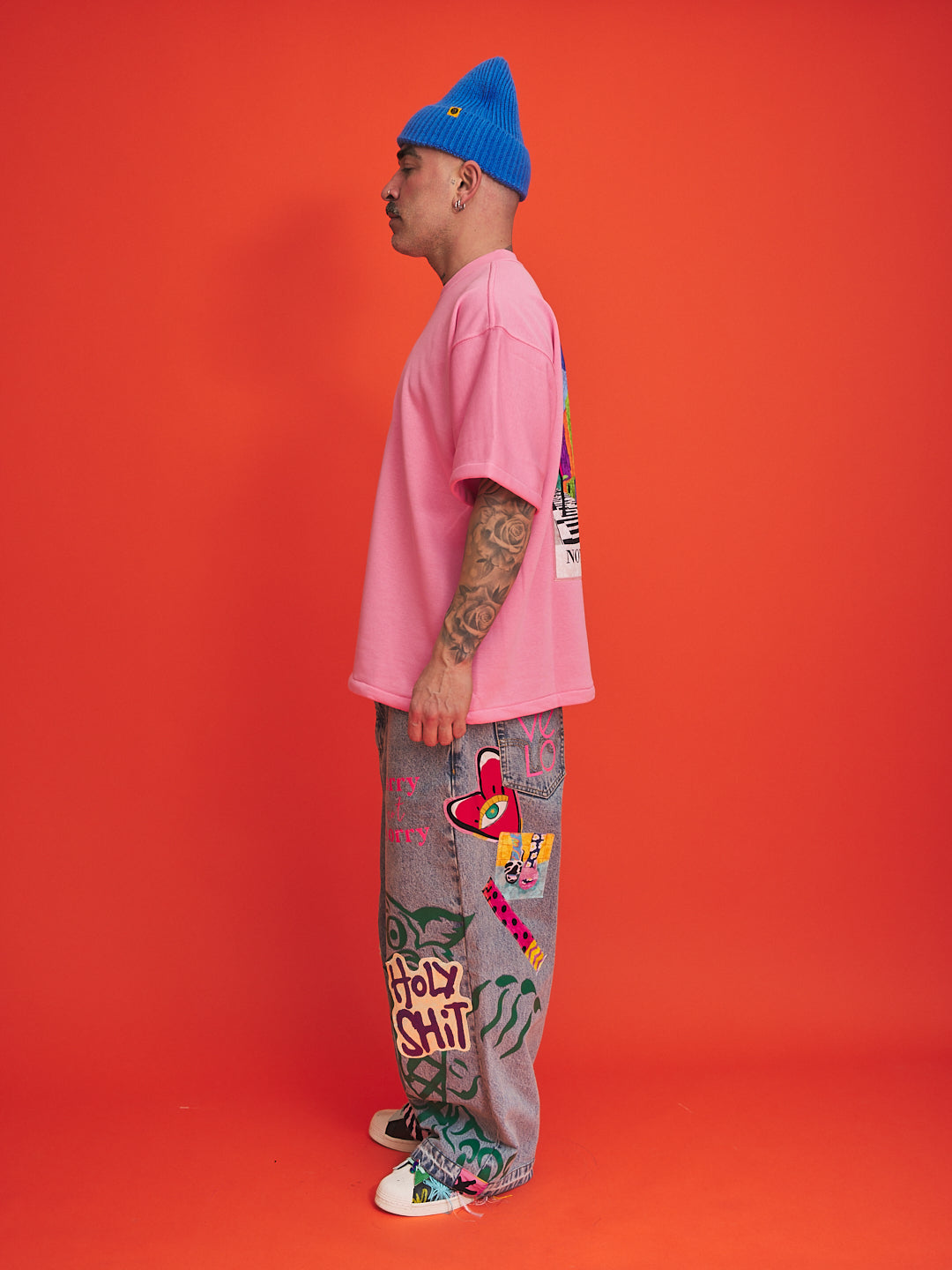 Person wearing a pink shirt, blue beanie, and gray pants with colorful designs on an orange background