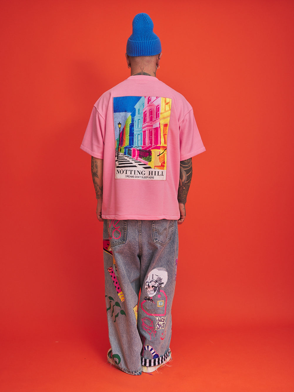 Person wearing a pink t-shirt with colorful graphic design and blue beanie on an orange background