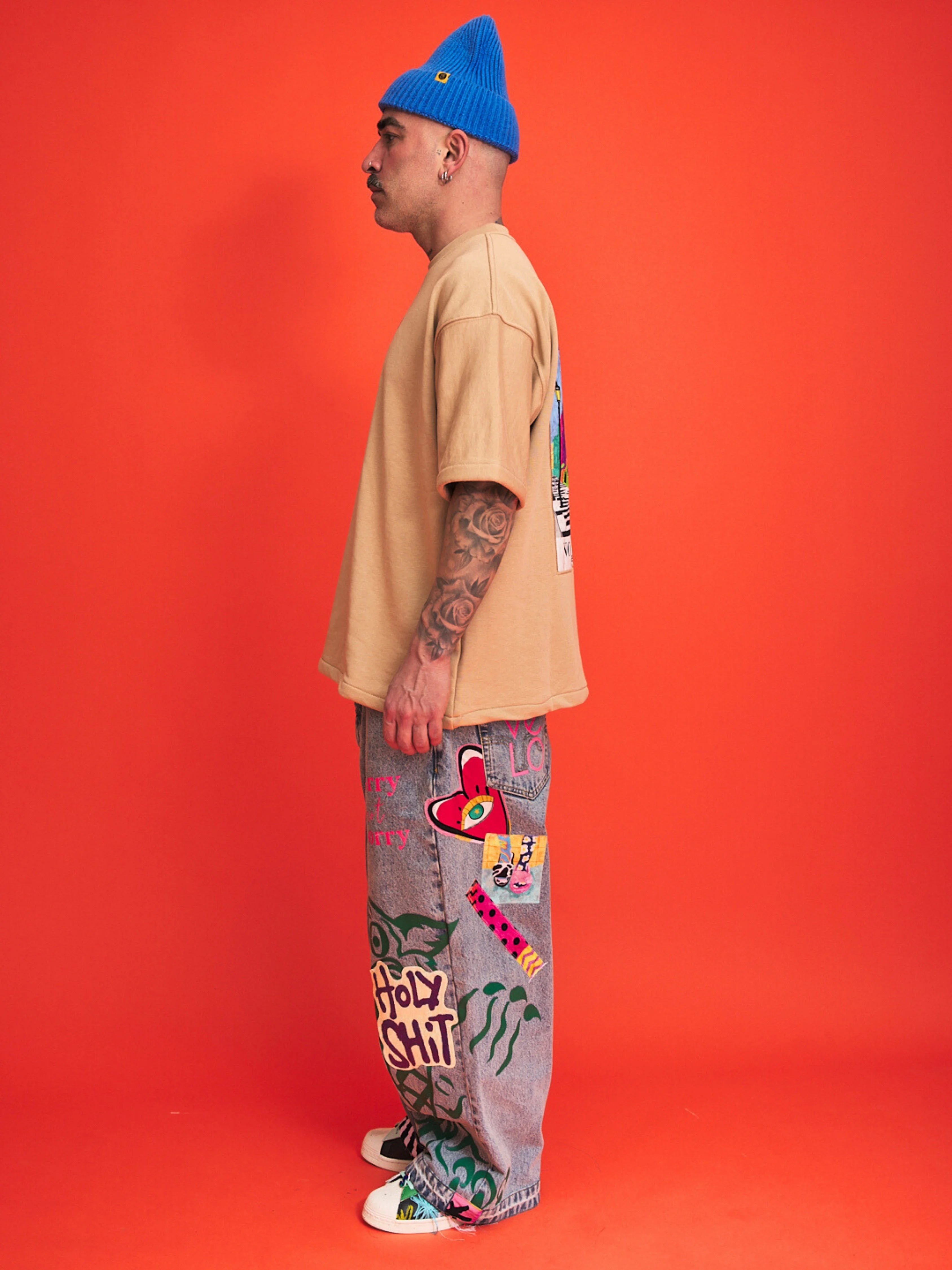 Person wearing a beige shirt, blue beanie, and colorful pants against an orange background