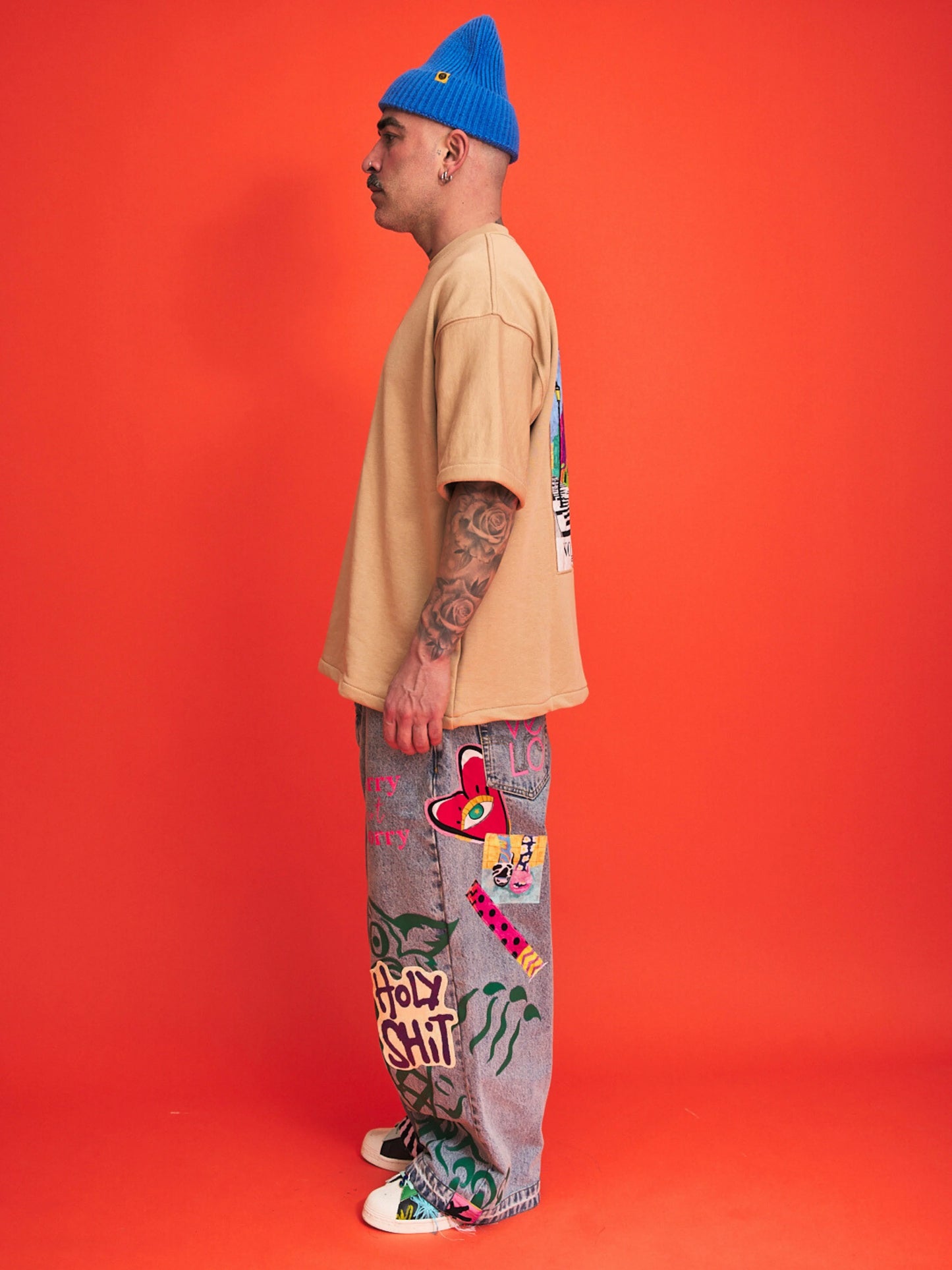 Person wearing a beige shirt, blue beanie, and colorful pants against an orange background